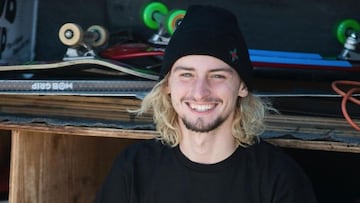 El skater Henry Gartland, con gorro negro y el pelo rubio saliéndole por detrás, así como unas tablas de skate a su espalda, mirando a cámara y sonriendo.