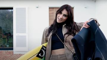 La surfsita Victoria Vergara sonríe con la cabeza de lado a la salida de su casa en Biarritz (Francia) con el neopreno en una mano y la tabla de surf en la otra, vestida a la moda para un shooting de Vogue.