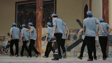Un mismo skater clonado varias veces con pantalones negros, tabla de skate, camiseta clara y gorra azul, andando frente a un spot en el que ha intentado un truco.