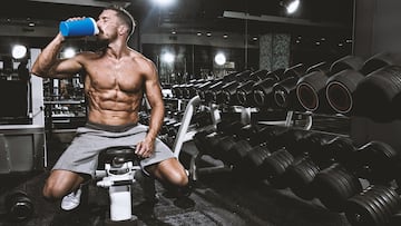 Nicely shaped male bodybuilder sitting on the bench next to dumbbells weight stand and drinking protein shake smoothy. There are mirrors on the walls.