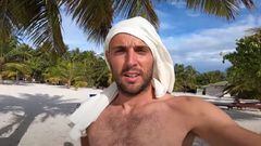 El surfista Borja Agote con una camiseta blanca en la cabeza para protegerse del sol en la playa de un resort de Maldivas, con palmeras al fondo y el cielo despejado con algunas nubes. 
