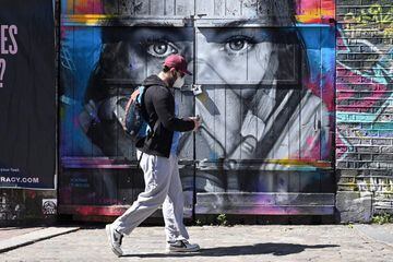 A man wearing a protective face mask walks past a piece of grafitti of artist BK Foxx wearing her grafitti mask created by French street artist Zabou in East London on April 19, 2020, during the novel coronavirus COVID-19 pandemic. 