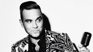 El cantante brit&aacute;nico Robbie Williams en una imagen en blanco y negro en la que aparece con una media sonrisa mirando un micr&oacute;fono que tiene en la mano y con un llamativo traje. 