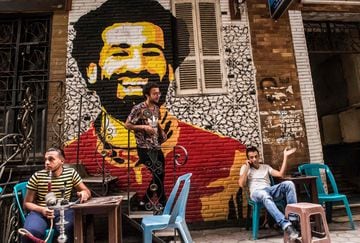 Ahmed Fathy, un artista egipcio de 26 años, desciende por una pequeña escalera de un café al aire libre en el centro de El Cairo. Tras él el mural que pintó del rostro del delantero egipcio del Liverpool, Mohamed Salah.