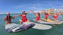Cinco surfistas con su tabla de surf cogi&eacute;ndose las manos y vestidas de Mam&aacute; Noel en la playa de Waikiki, Haw&aacute;i (Estados Unidos), el 25 de diciembre del 2022. 