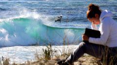 Un bodyboarder realiza un aéreo en una ola al fondo, mientras una chica en primer plano y desenfocada estudia de un libro.