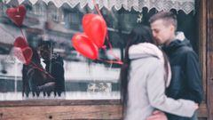 Una pareja bes&aacute;ndose frente a un escaparate mientras sujetan unos globos con forma de coraz&oacute;n