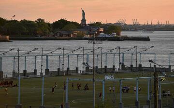 25. Gente jugando un partido de fútbol durante la puesta de sol en el Brooklyn Bridge Park en Nueva York con la Estatua de la Libertad al fondo.