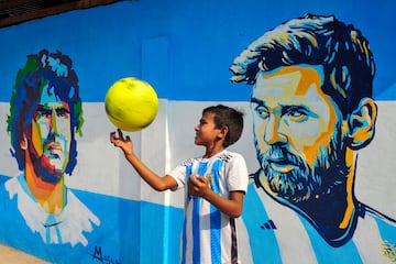 Un niño vestido con la camiseta de Argentina juega con una pelota frente a un mural en Bangladesh de las grandes leyendas de la selección albiceleste, Diego Armando Maradona y Lionel Messi, mientras los aficionados celebraban la victoria en las semifinales del Mundial de Qatar contra Croacia. 