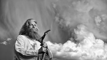 A black and white photo of an old man with long grey hair and a long grey beard standing in front of a dramatic dark sky. He is wearing a white toga and holding a wooden bar. He is looking up to the sky.