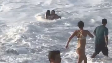 El surfista Mikey Wright, en espuma blanca generada por las olas en una playa de Haw&aacute;i (Estados Unidos), abraza a una chica a la que est&aacute; rescatando de las corrientes y el oleaje mientras, en la orilla y en bikini, su hermana Tyler Wright -b