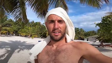 El surfista Borja Agote con una camiseta blanca en la cabeza para protegerse del sol en la playa de un resort de Maldivas, con palmeras al fondo y el cielo despejado con algunas nubes.