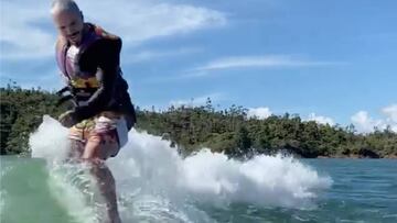 El cantante J Balvin, en bañador y con chaleco salvavidas, surfea una ola generada por el barco que va delante, con árboles de fondo.