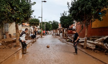 Dos chicos juegan al fútbol con botas de agua en una calle de Alfafar, mientras varias personas realizan labores de limpieza al fondo de la misma. Diez días después de la DANA, los servicios de emergencia apuran hasta el límite la búsqueda de desaparecidos. Las víctimas mortales se mantienen en 219 y los desaparecidos, por el momento, son 78.