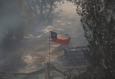 La bandera nacional de Chile ondea en medio del humo y cerca de los restos quemados de una casa.