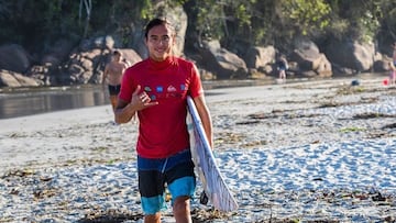El surfista brasileño Pedro Tanaka, con la playa y unas rocas de fondo y vestido con un bañador corto de líneas azules y negras y una licra roja, con la tabla de surf bajo el brazo, realiza el señal surfero del shaka mientras s