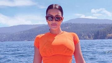 La modelo Kim Kardashian vestida de naranja y con gafas de sol en la punta de un yate en un lago, con monta&ntilde;as al fondo. 