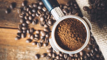 Freshly ground espresso in a portafilter, ready to be made into a hot shot of espresso. Rustic wood background, a burlap coffee bag in the background covered with roasted coffee beans. Overhead view.