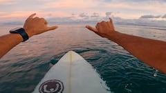 Parker Seidel hace el shaka con las dos manos sobre su tabla de surf en el agua. 