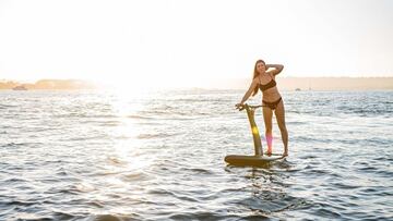 Una chica en bañador surfea con un hydrofoiler por un mar en calma, con el sol quemando la imagen al fondo.