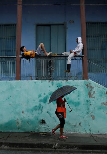Una mujer con paraguas pasa junto a dos personas en un porche antes de la llegada del huracán Melissa, en Santiago de Cuba, Cuba.