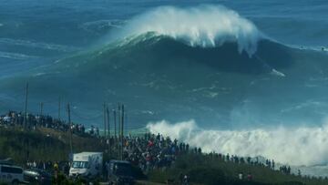 La surfista francesa Justine Dupont surfea una ola gigante en Nazaré (Portugal) con el acantilado en primer plano lleno de gente y furgonetas observando la sesión de surf de olas grandes.