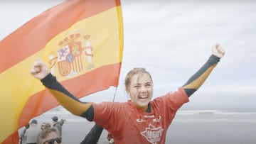 Carmen L&oacute;pez saliendo a hombros del agua en La Jolla (California, Estados Unidos) el 14 de marzo del 2020, levantando los brazos en se&ntilde;al de victoria y sonriendo, con una bandera espa&ntilde;ola detr&aacute;s y unas cuantas olas al fondo de 