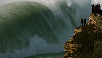 Una ola gigante rompiendo frente al faro de Praia do Norte, Nazar&eacute; (Portugal), con espectadores mirando desde las rocas. 