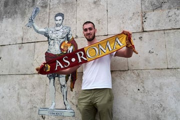 Un aficionado de la Roma sostiene la bufanda de su equipo ante una pintura del artista Harry Greb que representa al entrenador de la Roma, José Mourinho, caracterizado como un emperador romano que levanta victorioso el trofeo de campeón de la Conference League conquistada el miércoles ante el Feyenoord en Tirana.