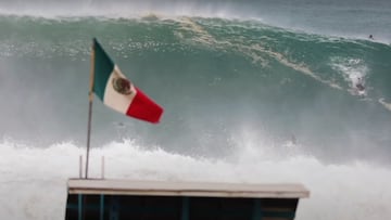 Una ola gigante a punto de romper en la playa de Zicatela (M&eacute;xico), con la bandera del pa&iacute;s ondeando en primer plano sobre la caseta dle socorrista y un surfista intentando hacer el drop a la derecha. 