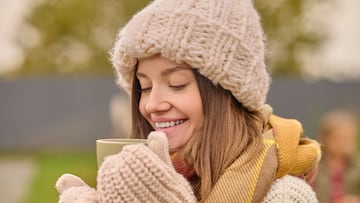 Pleasure. Young beautiful woman in knitted hat and mittens looking with pleasure into cup with hot drink standing outdoors on cool autumn day