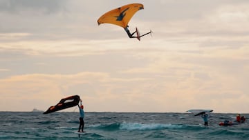 Un chico practicando wing foil en Tarifa (C&aacute;diz, Espa&ntilde;a) con una cometa de F-One, volando muy alto, mientras otro levanta los brazos en se&ntilde;al de victoria al verle pasar por encima. 