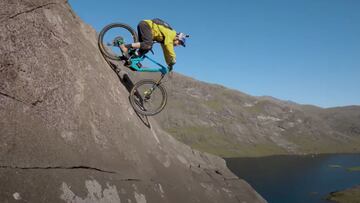 Danny MacAskill bajando con su bici por las rocas de Dubh Slabs, en la isla de Skye (Escocia) con su bici de MTB Santa Cruz azul, ropa de color amarillo, el casco Red Bull y la GoPro. El lago Coruisk, al fondo. 