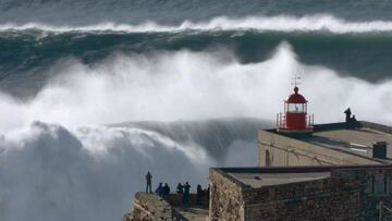 Pedro Scooby a punto de entrar en una ola en Nazaré (Portugal) durante una gran marejada en Praia do Norte. Al fondo se ven olas gigantes formándose y rompiendo. En primer plano, el faro de San Miguel Arcángel.