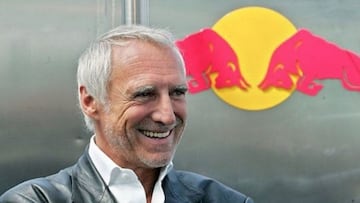 Dietrich Mateschitz sonriendo, vestido con una americana de cuero, frente a un logo de la compa&ntilde;&iacute;a Red Bull sobre fondo plateado. 