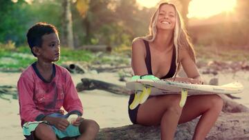 Surfista francesa Maud Le Car en bikini negro y con su tabla de surf en las Mentawai, sentada al lado de un niño local.