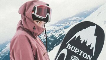 La snowboarder Maria Hidalgo mirando a cámara tapada con máscara, gorro y chaqueta, mostrando también su tabla de snowboard en la montaña cubierta de nieve.
