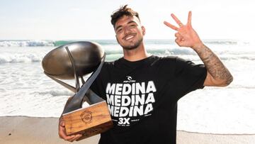 Gabriel Medina levanta 3 dedos en relación a sus tres campeonatos del mundo de surf de la WSL, con el trofeo en la otra mano, una camiseta negra en la que se puede leer tres veces Medina y, de fondo, la ola de Lower Trestles, en San Clemente (California,
