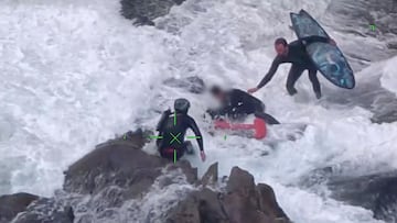Dos miembros del equipo de rescate de la Policía de Nueva Zelanda en el rescate a un surfista entre las rocas de una playa de arena negra.