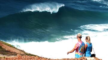 La ola gigante de Nazar&eacute; a punto de romper, vista desde el acantilado del pueblo portugu&eacute;s, con algunos espectadores andando por la zona.