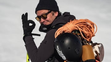 Xavier De Le Rue toc&aacute;ndose el dedo pulgar con el &iacute;ndice y mirando a c&aacute;mara con gafas de sol, gorro y equipamiento para la aventura como una cuerda y un casco, en un paisaje que se puede ver al fondo cargado de nieve. 