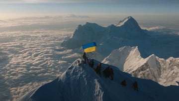 Tonya Samoilova, con la bandera de Ucrania, en el Everest, grabada en dron, con nubes debajo y el sol al fondo.