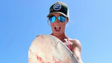 La patrona de barco Kaylee Crisp posa con un tibur&oacute;n pescado por ella en su barco, abriendo la boca, con gafas de sol y gorra, con un despejado cielo azul de fondo. 
