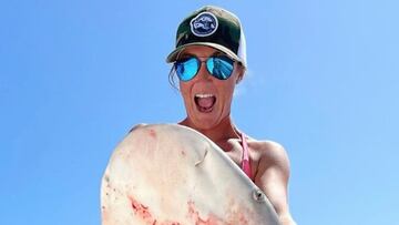 La patrona de barco Kaylee Crisp posa con un tiburón pescado por ella en su barco, abriendo la boca, con gafas de sol y gorra, con un despejado cielo azul de fondo.