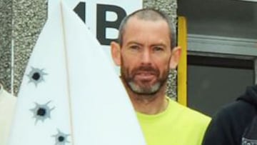 El surfista brit&aacute;nico Lee Bartlett, con una camiseta amarilla y rapado, mira a c&aacute;mara con una tabla de surf con pegatinas de disparos. 