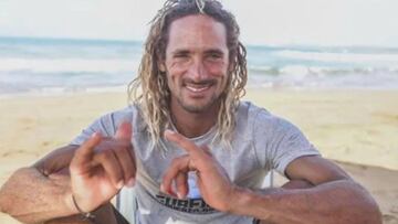 Brian Ramos Torres, surfista de Puerto Rico, en la playa con el mar de fondo sonriendo y haciendo el símbolo surfero del Shaka. En la camiseta se puede leer Surfing Puerto Rico, su escuela de surf de la playa de La Pared, en Luquillo.
