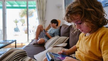 Boy playing on a digital tablet on the sofa at home, with his brother in the background