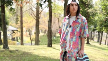 El skater Isamu Yamamoto posando con una camisa de colores y su tabla de skate, con gorra blanca, frente a unos árboles.