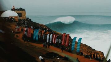 Una ola gigante rompe en la Praia do Norte de Nazare; frente al acantilado de San Miguel Arcangel, con muchas banderolas de un evento de surf.
