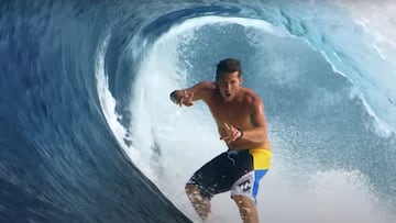 El triple campeón del mundo de surf Andy Irons (1978 - 2010) sin camiseta y en bañador Billabong surfeando una ola en forma de tubo -de derechas- y haciendo el símbolo de la victoria con ambas manos mientras mira a cámara.